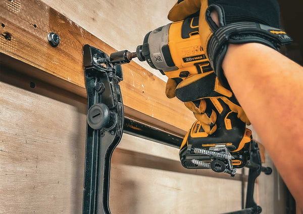 A DeWalt impact driver is being used to insert lag screws into a wooden stud, securing a TV mount. The Second Set magnet attached to the impact driver is holding onto additional lag screws for easy access. The person operating the tool is wearing protective gloves.