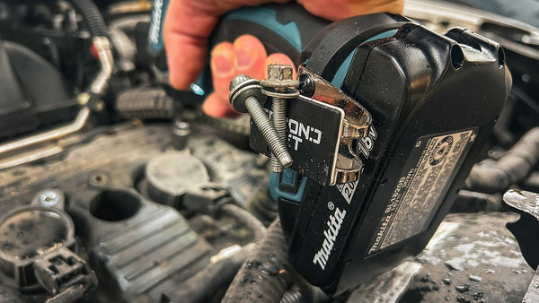 A close-up of a Makita drill in use under a car hood, equipped with a magnetic attachment securely holding bolts, demonstrating its practical application for engine repairs.